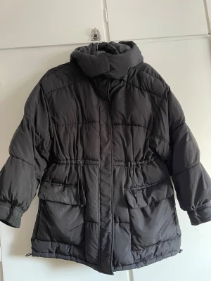 Svart pufferjacka från NA-KD - Svart pufferjacka från NA-KD med hög krage, dragsko i midjan och stora fickor framtill. Jackan har en oversized passform och är perfekt för kalla vinterdagar. Tillverkad i syntetmaterial och har en stilren look som passar till det mesta.