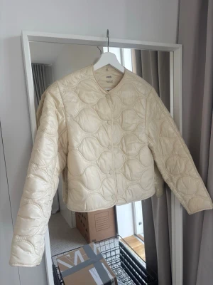 Krämvit pufferjacka från Mango - Populära pufferjackan från Mango med vågigt quiltat mönster och rundad halsringning. Jackan är kortare i modellen och har tryckknappar framtill. Perfekt nu i vår för att hålla värmen och samtidigt se trendig ut. Storlek xxs men passar även en xs.