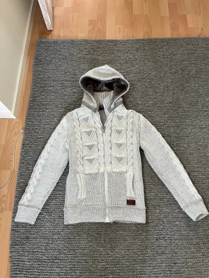 Stickad grå hoodie med dragkedja - Snygg stickad hoodie i ljusgrått med dragkedja framtill och kabelstickat mönster på ärmar och framstycke. Fodrad huva för extra värme och två fickor framtill. Perfekt för dig som gillar en mysig och stilren look.