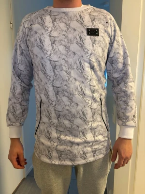 Vit sweatshirt med marmormönster criminal damage - Säljer en vit sweatshirt från criminal damage med grått marmormönster. Tröjan har svarta dragkedjor på fickorna och en svart logopatch på bröstet. Ribbstickade muddar vid ärmslut och hals. Perfekt för dig som gillar streetwear och unik design. Storlek L, finns litet hål på vänster armhåla annars bra 