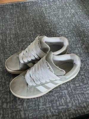 Adidas gråvita sneakers i mocka - Snygga Adidas sneakers i grå mocka med klassiska vita sopor och vit sula. Skorna har lågt skaft och breda vita skosnören. Perfekta för dig som gillar en clean och sportig look. Ikonisk Adidas-logga på, och bekväm vadderad sopkant.
