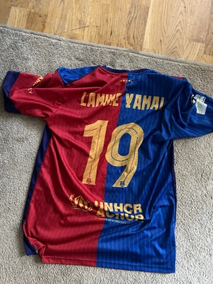FC Barcelona pass touching tröja Nike - Säljer en FC Barcelona matchtröja med Lamine Yamal och nummer 19 på ryggen. Tröjan är r folding i rött och blått med gula detaljer, klubbmärke och Nike-logga på bröstet samt Spotify-logga. Tillverkad i lätt och ventilerande material, perfekt för fotboll eller träning.