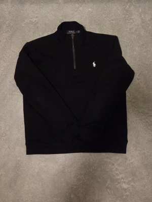 Svart half-zip tröja från Polo Ralph Lauren - Klassisk svart långärmad tröja från Polo Ralph Lauren med half-zip och hög krage. Tröjan har den ikoniska vita logon broderad på bröstet och är tillverkad i mjukt material. Perfekt för en stilren och avslappnad look. Storlek L enligt lappen, men passar bäst som S.