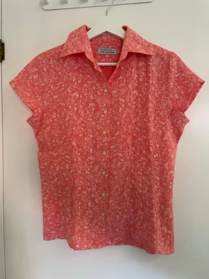Blommig rosa skjorta  - Kortärmad skjorta från The shorts factory (design Linnéa Braun) i en härlig rosa färg med små vita blommor. Skjortan har klassisk krage, knappar framtill och är gjord i mjuk bomull. Perfekt för dig som gillar en söt och avslappnad stil. Använt 2 gånger!