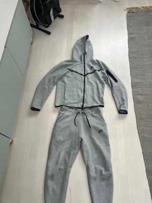 Grå Nike Tech mjukisdress med huva - Säljer en grå mjukisdress från Nike med dragkedja på tröjan och huva. Byxorna fickor med dragkedja. Setet har svarta detaljer och Nike-logga på både tröja och byxa. Perfekt för chill dagar eller träning.