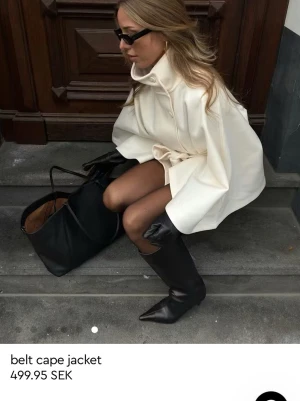 Krämvit puncho-kappa med bälte - Helt ny pris lapp finns på , Snygg cape-kappa i krämvit färg med hög krage och knäppning framtill. Kappan har ett matchande bälte i midjan som ger en cool och modern siluett. Materialet ser ut att vara mjukt och lite tjockare, perfekt för kyliga dagar.