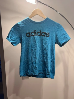Blå Adidas t-shirt med logga - Snygg blå t-shirt från Adidas med stor svart logga över bröstet. Klassisk rund halsringning och korta ärmar. Perfekt för en sportig och avslappnad stil. Tillverkad i mjuk bomull för skön känsla.