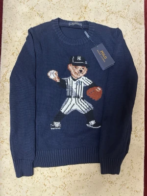 Mörkblå stickad tröja Polo Ralph Lauren - Mörkblå stickad tröja från Polo Ralph Lauren med ett broderat motiv av en basebollspelare på framsidan. Tröjan har rund halsringning och ribbade muddar vid ärmslut och nederkant. Perfekt för dig som gillar sportiga detaljer och klassisk stil.