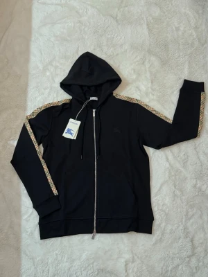 Svart hoodie från Burberry med dragkedja - Snygg svart hoodie från Burberry med dragkedja framtill och klassiskt Burberry-mönster längs ärmarna. 