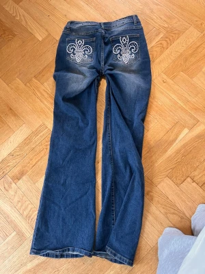 Blå bootcut jeans med brodyr - Säljer ett par blå bootcut jeans med vita broderade mönster på bakfickorna. Jeansen har klassisk femficksdesign och lätt slitna detaljer framtill. Perfekta för dig som gillar en avslappnad men ändå unik stil. Jag har sytt dem om till low rise men det går lätt att sprätta upp då jag inte klippt bort något tyg, materialet är ganska strechigt 💕 fråga om mått! 💝