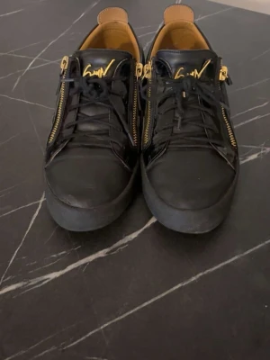  Giuseppe Zanotti skor svarta äkta  - Snygga svarta sneakers i läder från Giuseppe Zanotti med guldfärgade dragkedjor på sidorna och guldfärgad logga på plösen. Skorna har svarta snören och en chunky sula. Insidan är ljusbrun och materialet känns lyxigt och stilrent.