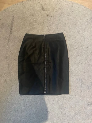 Svart kjol med dragkedja framtill - Snygg svart skinn kortkjol med en markerad dragkedja som går längs hela framsidan. Kjolen har en rak passform och är enkel att matcha med olika stilar. Perfekt för dig som gillar minimalistiska och stilrena plagg.