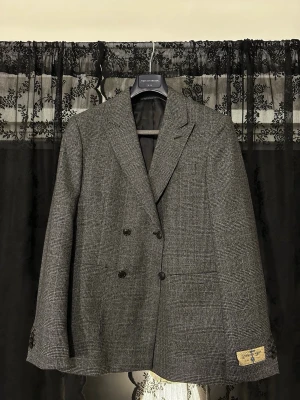 Grå dubbelknäppt kavaj These Glory Days  - Nypris 4999 kr. HELT OANVÄND! Snygg grå kavaj från These Glory Days med klassiskt rutmönster och dubbelknäppning i storlek 48. Kavajen har spetsiga slag, fyra knappar vid ärmslut. Perfekt för dig som gillar stilrena och tidlösa plagg.