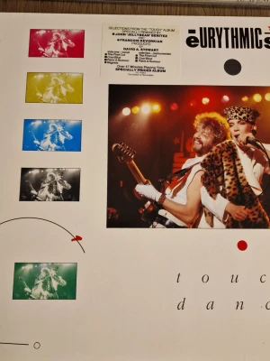 Eurythmics Touch Dance vinylskiva - Säljer en vinylskiva med albumet 'Touch Dance' av Eurythmics. Omslaget är vitt med färgglada foton och grafiska detaljer, och innehåller information om låtar och medverkande. Perfekt för dig som gillar synthpop och 80-talsmusik.