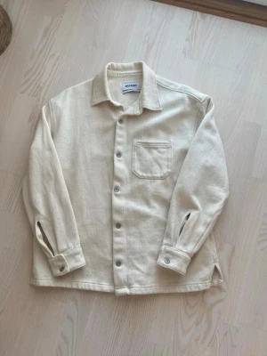 Overshirt från Weekday - Stilren beige overshirt från Weekday i kraftigt bomullstyg. Klassisk krage, knappstängning framtill och en bröstficka. Perfekt lager-på-lager-plagg med avslappnad passform och diskreta detaljer.
