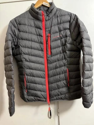 Grå dunjacka från High mountain  - Säljer en grå dunjacka från  High mountain  med röda dragkedjor och quiltad design. Jackan har hög krage, tre fickor med dragkedja och lätt vaddering för extra värme. Perfekt för kalla vinterdagar och outdoor-äventyr.