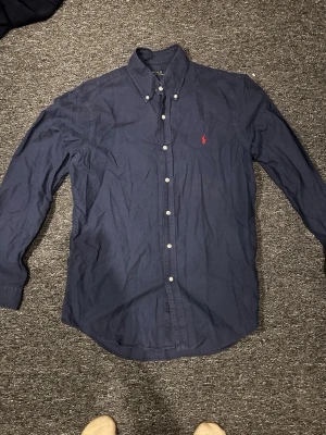 Marinblå skjorta från Ralph Lauren - Klassisk marinblå skjorta från Ralph Lauren med röd broderad logga på bröstet. Skjortan har knappar hela vägen, button-down krage och långa ärmar. Perfekt till jeans eller chinos för en clean och stilren look.