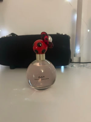 Dot parfym från Marc Jacobs - Säljer en Dot parfym från Marc Jacobs. Flaskan är rund och genomskinlig med ljusrosa doftvätska. Locket är rött med svarta prickar och dekorerat med en blomma och gulddetaljer, vilket ger en lekfull och unik look. Perfekt för dig som gillar snygg design på parfymflaskan. Endast testad 