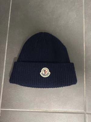 Mörkblå ribbstickad Moncler mössa - Säljer en mörkblå ribbstickad mössa från Moncler med uppvikt blind och klassisk Moncler-logga framtill. Mössan är i mjukt material och har en enkel, stilren design som passar perfekt till kalla dagar.