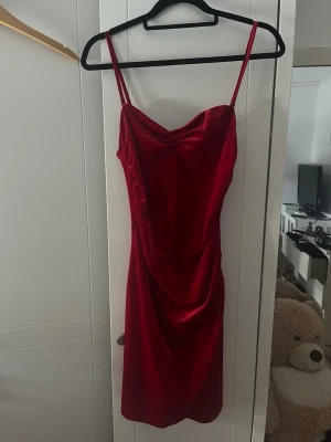 Röd sammetsklänning med smala band - Snygg röd klänning i mjuk sammet med smala axelband och draperad urringning. Kort modell som ger en elegant siluett och passar perfekt för dig som vill sticka ut. Enkel men ändå lyxig känsla tack vare materialet.