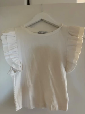 Vit ribbad topp med volangärm Zara - Supersöt vit ribbad topp från Zara med volangdetaljer på ärmarna. Toppen har rund halsringning och är tillverkad i ett mjukt bomullsmaterial. Perfekt för dig som gillar en enkel men ändå trendig look.
