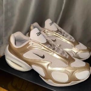 Nike air max muse beige/guld/vit - Nike Air max muse sneakers i metallic guld och vitt med meshpaneler och  detaljer. Låg modell med svart swoosh på sidan och chunky sula med synlig dämpning. Perfekta för dig season som vill sticka ut med en unik och sportig look.