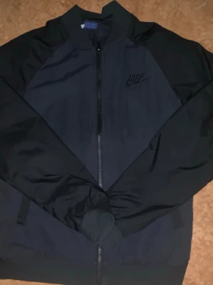 Svart vindjacka från Nike - Snygg svart vindjacka från Nike med dragkedja framtill och diskret broderad logga på bröstet. Jackan har ribbade muddar och är tillverkad i ett lätt, vindtätt material som passar perfekt till streetwear och sportiga outfits.