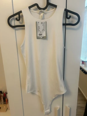 Body H&M - Säljer en vit bodysuit från H&M med rund halsringning och ärmlös design. Bodyn har en tight passform och är tillverkad i mjuk bomull, perfekt för att skapa en clean och enkel look.