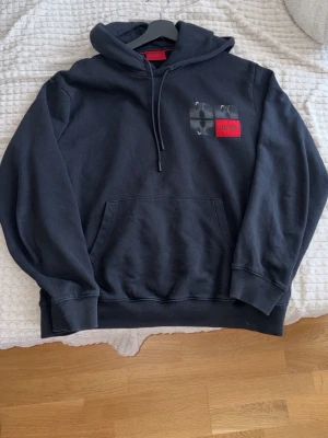 Svart hoodie från Hugo med tryck - Snygg svart hoodie från Hugo med stor grafisk print på ryggen och röd logotyp på bröstet. Klassisk känguruficka framtill, ribbade muddar sopch huva med. Tillverkad i mjuk bomullsblandning för skön känsla. Perfekt för en streetwear-look. Hål vid fickan (se tredje bild) men syns ej när man har på sig den. Kommer från ett djur och rökfritt hem.