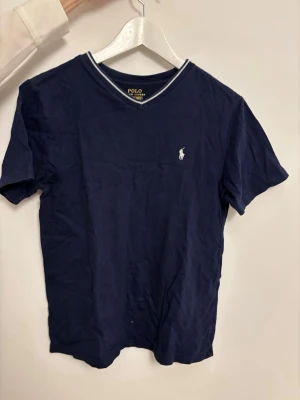 Marinblå t-shirt från Polo Ralph Lauren - Klassisk marinblå t-shirt från Polo Ralph Lauren med vit broderad logga på bröstet och v-ringad hals med vit kant. Tillverkad i mjuk bomull och har korta ärmar. Perfekt basplagg för en clean och stilren look.🤩
