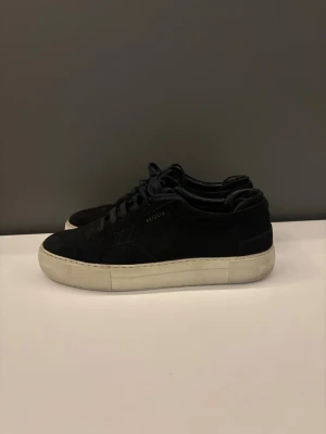 Svarta sneakers från Axel Arigato - Stilrena svarta sneakers från Axel Arigato med ovandel i mocka och klassisk vit sula. Låga i modellen med svarta skosnören och diskret logga på sidan. Perfekta för en clean och modern look. Ett av skosnörehålen har gått sönder men funkar ändå att använda. Ett litet svart märke finns även på den vita sulan på den ena skon men inget som märks särskilt mycket. Är i storlek 42