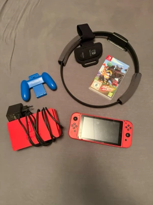 Nintendo Switch med Ring Fit Adventure - Nintendo Switch-konsol i rött med Ring Fit Adventure, ring och benrem, blå Joy-Con-hållare, röd dockningsstation och laddare. Perfekt för gaming och träning. Allt du behöver för att spela direkt och utmana dig själv med roliga spel. Kommer inte med nån laddare