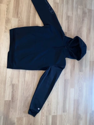 Svart hoodie från Champion - En klassisk svart hoodie från Champion med huva och känguruficka framtill. Diskret Champion-logga på ena ärmen och dragsko i huvan. Tillverkad i mjukt bomullsmaterial som är skönt mot huden. Perfekt för en avslappnad och sportig stil.