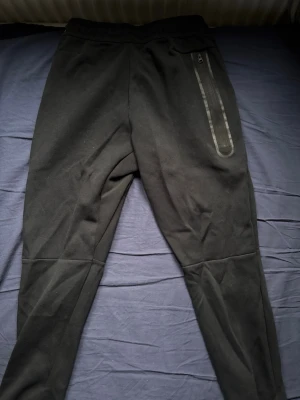 Svarta Nike Tech Byxor - Svarta mjukisbyxor från Nike med modern passform och dragkedjeficka på höger sida. Byxorna har elastisk midja och är tillverkade i mjukt material som är perfekt för chill eller träning. Snygga detaljer vid knäna ger extra stil.