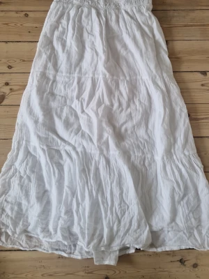 Vit långkjol från Gina Tricot - Säljer en vit långkjol från Gina Tricot med smock i midjan och lätt rynkad struktur. Kjolen är tillverkad i ett tunt och luftigt bomullsmaterial, perfekt för varma dagar. Enkel och stilren design som är lätt att matcha med olika toppar.