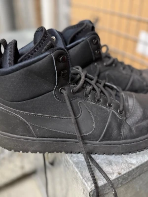 Svarta Nike high top sneakers - Säljer ett par helsvarta high top sneakers från Nike med klassisk swoosh på sidan. Skorna har snörning, slitstark yttersula och är tillverkade i läder och syntetmaterial. Perfekta för dig som gillar en clean och sportig look.