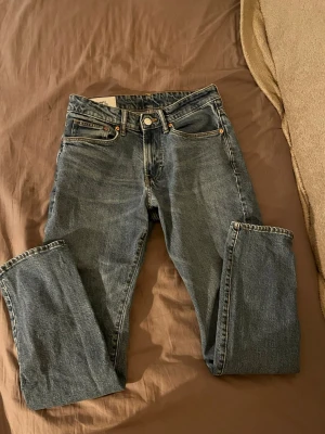 Blå raka jeans från H&M - Klassiska blå jeans från H&M med straight regular fit 29/32 och fem fickor. Jeansen har en tidlös tvätt och mässing färgade nitar. Tillverkade i slitstarkt denimtyg med knappgylf och bälteshällor. Perfekta för en avslappnad och stilren look.