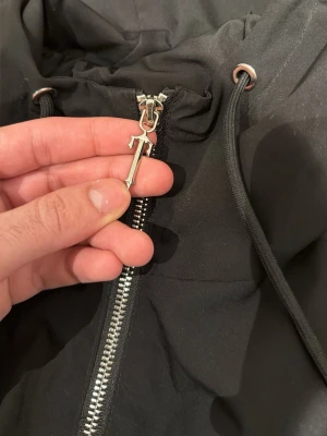 Svart vindjacka från Trapstar - Säljer en svart vindjacka från Trapstar med stor logga broderad på ryggen och ikonisk T-dragkedja. Jackan har huva med snörning och en patch på ärmen med texten 'It's a secret'. Perfekt för dig som vill ha streetwear-vibe.