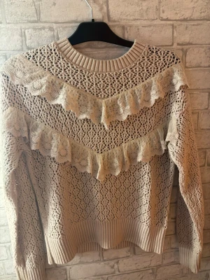 Lindex  - Supersöt beige stickad tröja med genombrutet mönster och två breda spetsvolanger över bröstet och armarna. Tröjan har rund halsringning, ribbade muddar och är perfekt för dig som gillar romantisk och bohemisk stil. Materialet känns mjukt och luftigt.