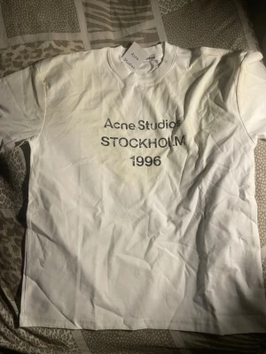 Vit Acne Studios t-shirt med tryck - Säljer en vit t-shirt från Acne Studios med svart texttryck på bröstet där det står 'Acne Studios STOCKHOLM 1996'. Klassisk rund hals och korta ärmar. Tillverkad i mjuk bomull för en clean och stilren look. Kan ge bra pris till rätt person 