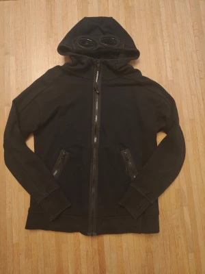 C.P. Company Hoodie. Perfekt skick. Nypris 2990kr - Använd två gånger men säljes för den är för liten för mig, då jag har köpt en L istället. Nypris 2990kr på careofcarl. https://www.careofcarl.se/sv/cp-company-diagonal-raised-fleece-full-zip-goggle-hoodie-black-5?_gl Den är i perfekt skick som du kan se på bilderna. Tröja från C.P. Company med hel dragkedja och två fickor med dragkedja framtill. Huvan har inbyggda glasögon som ger en unik och cool look. Tillverkad i mjukt bomullsmaterial och har ribbade muddar vid ärmar och nederkant.