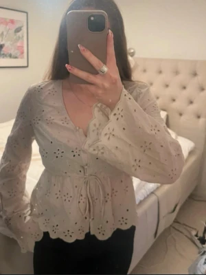 Beige broderad blus med knytband - Söt beige blus med broderade blommor och hålmönster. Blusen har v-ringning, volangkant och knytband i midjan. Ärmarna är långa och vida med vågig avslutning. Perfekt för en romantisk och trendig look.