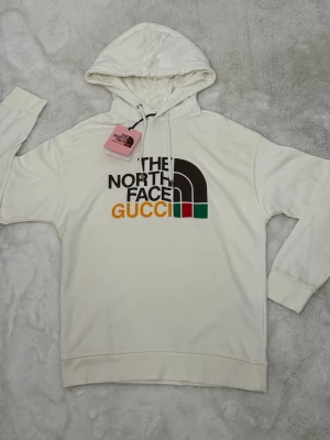 Vit hoodie The North Face x Gucci - Snygg vit hoodie med huva från The North Face x Gucci. Stor logga framtill i svart, brunt, gult, grönt och rött. 