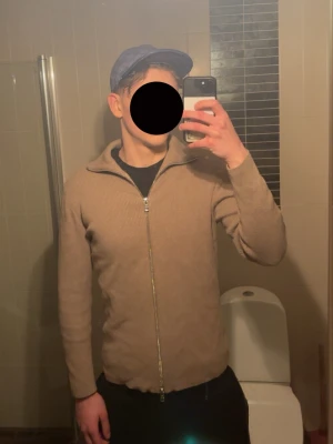 Beige ribbstickad ziptröja - Snygg beige ribbstickad tröja med hög krage och hel dragkedja framtill. Tröjan har lång ärm och är tillverkad i ett mjukt, stretchigt material som ger en clean och stilren look. Perfekt att slänga på sig över en t-shirt för extra värme.