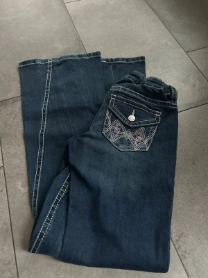 Mörkblå bootcut jeans med brodyr från Gina tricot  - Säljer ett par mörkblå bootcut jeans med dekorativa bakfickor som har vit brodyr och knappdetaljer. Jeansen har kontrastsömmar och klassisk femficksdesign. Perfekta för dig som gillar mer utsvängda ben och snygga detaljer. Från Gina tricot 