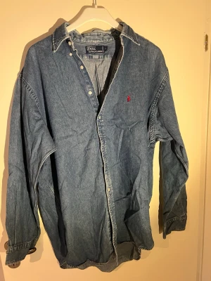 Blå jeansskjorta från Polo Ralph Lauren - Klassisk blå jeansskjorta från Polo Ralph Lauren med trust off röd broderad logga på bröstet. Skjortan har långa ärmar,?knappar framtill och en relaxed passform. Perfekt för en avslappnad och cool stil}]
