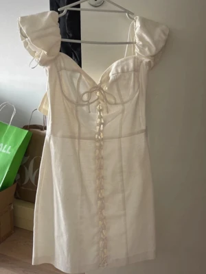 Vit kort klänning med puffärmar - Supersöt vit kort klänning från Sisters Point med puffiga kortärmar och snörning framtill. Klänningen har en hjärtformad urringning och är tillverkad i ett lätt tyg som passar perfekt till varmare dagar. Snygg och trendig modell med romantiska detaljer.