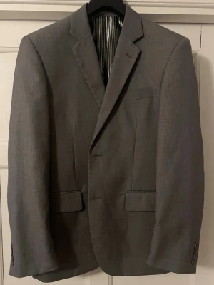 Grå kavaj från Everyday - Stilren grå kavaj från Everyday med klassisk passform och två knappar framtill. Kavajen har två fickor med lock och en bröstficka. Insidan är fodrad med grå- och vitrandigt tyg. Perfekt för en snygg och enkel look.