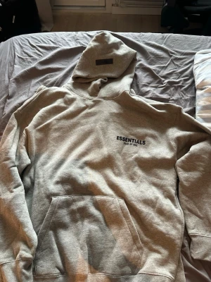 Grå Essentials Fear of God set - Säljer ett grått loungewear-set från Essentials Fear of God. Setet består av en hoodie med känguruficka och tryck på bröstet samt matchande mjukisbyxor med dragsko och tryck. Perfekt för chill dagar hemma. Materialet är mjukt och bekvämt bomullsmix.