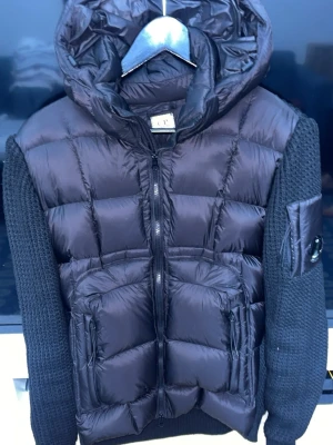 Svart hybrid-cardigan från C.P. Company - Snygg svart cardiganjacka från C.P. Company med vadderad framsida och huva i glansigt material samt stickade ärmar och rygg. Jackan har två dragkedjefickor fram och en ikonisk linsdetalj på ärmen. Perfekt för lager-på-lager-stil.